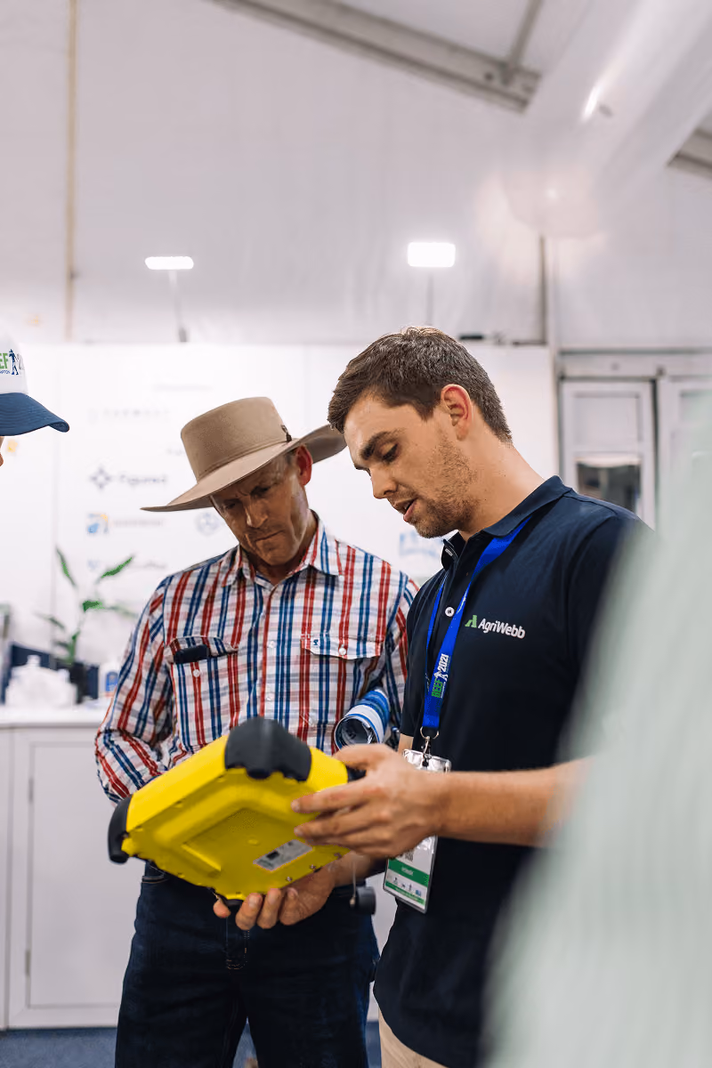 A young man in an AgriWebb shirt explaining a yellow handheld device to an older man wearing a wide-brimmed hat and plaid shirt inside a well-lit indoor setting.
