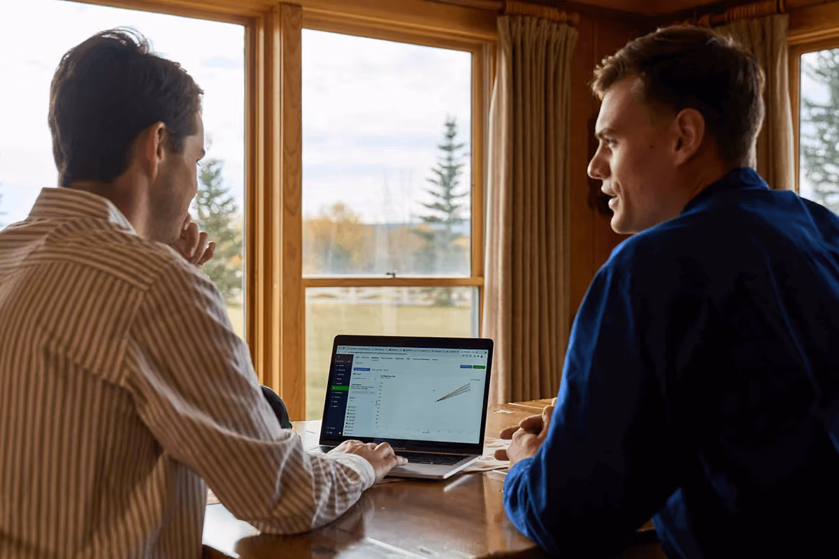 Two men sitting at a wooden table in a room with large windows, one man facing away from the camera and the other facing sideways, looking at a laptop displaying a chart on the screen, with a scenic outdoor view of trees and sky in the background.