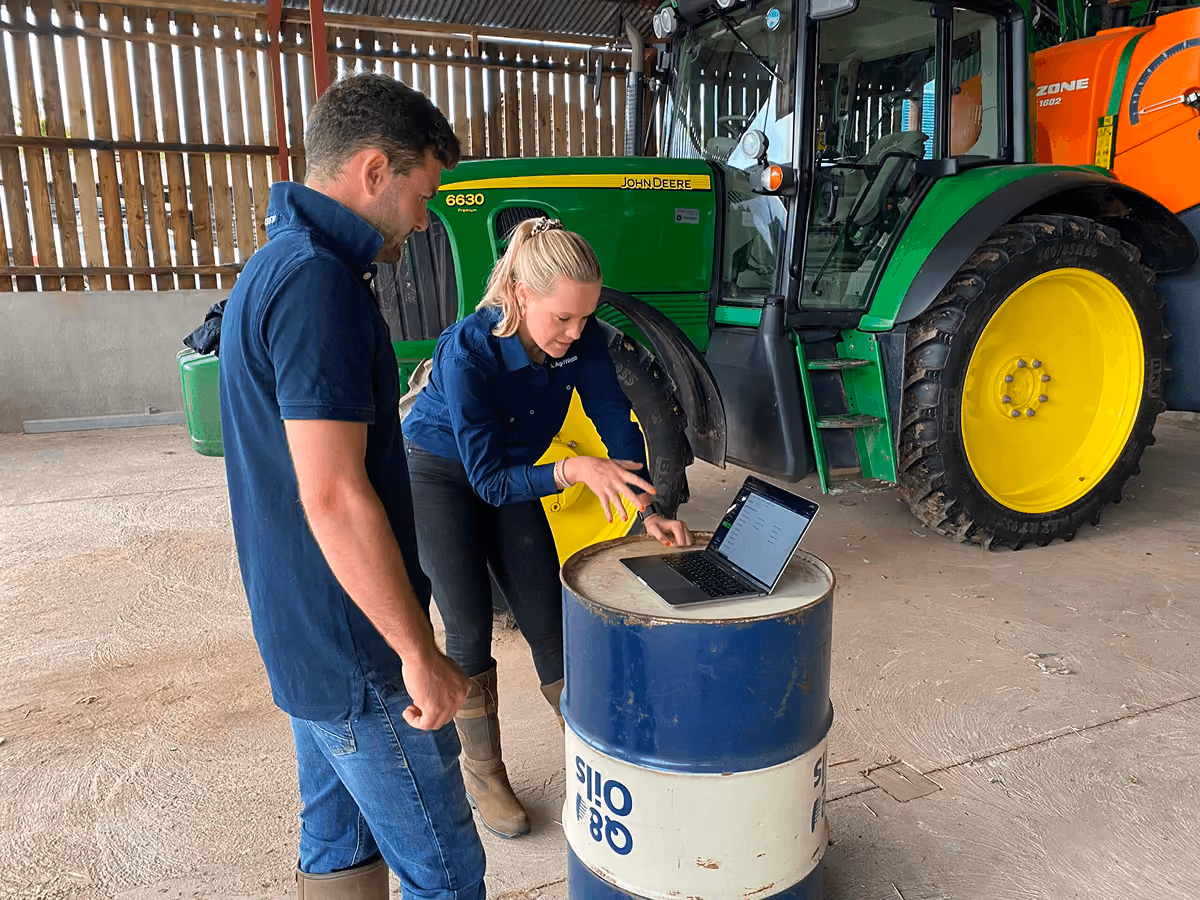 Two people in a barn or shed standing next to a green John Deere tractor, looking at a laptop placed on top of a blue and white metal barrel.