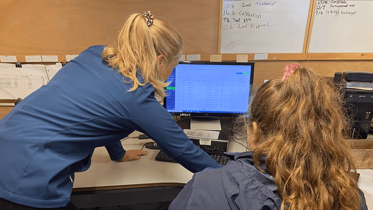 Two women with long hair in blue jackets working together at a desk while looking at a computer screen displaying data in an office setting.