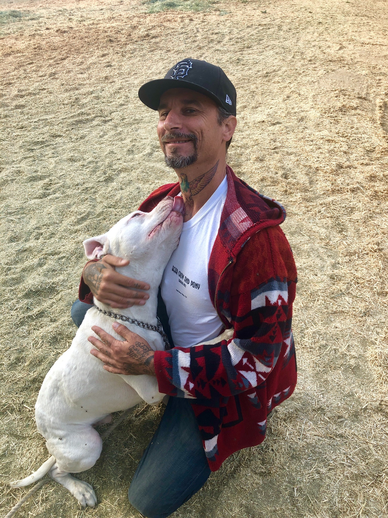 Greg Carroll kneeling outdoors while Ozzy, a white pit bull, affectionately leans against him.