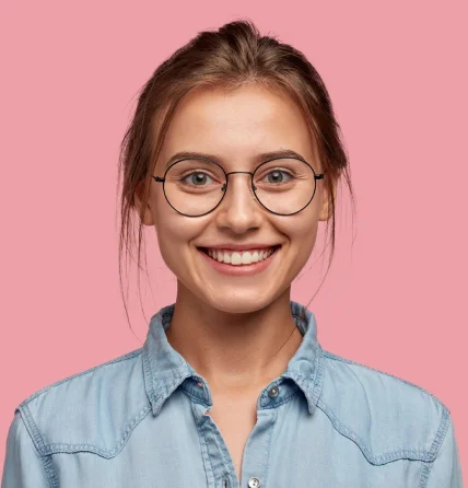 Young woman with light brown hair in a loose bun, wearing round eyeglasses and a light blue denim shirt, smiling against a pink background.