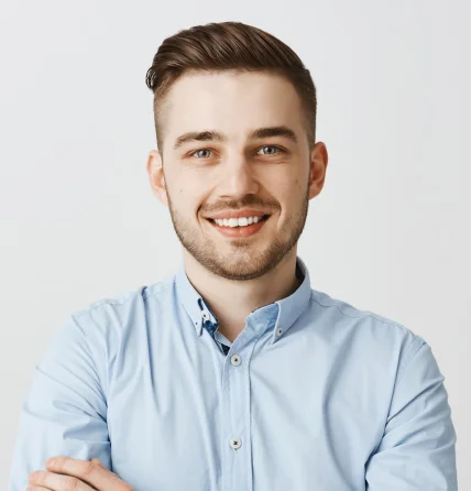 Smiling young man with short brown hair and beard, wearing a light blue button-up shirt, standing with arms crossed against a plain light background.