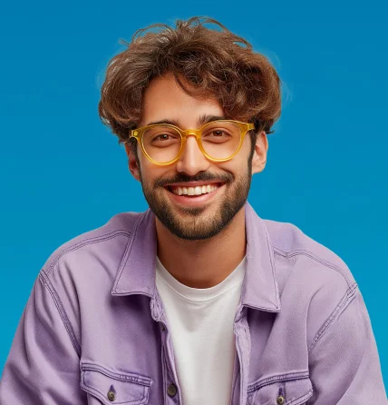 Smiling young man with curly brown hair, beard, and round yellow glasses wearing a lavender jacket over a white shirt against a blue background.