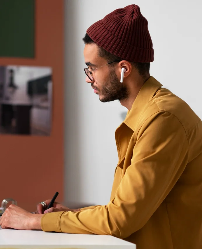 Man wearing a maroon knit hat, glasses, and a mustard yellow jacket, using a stylus to write or draw on a surface while wearing white wireless earbuds.