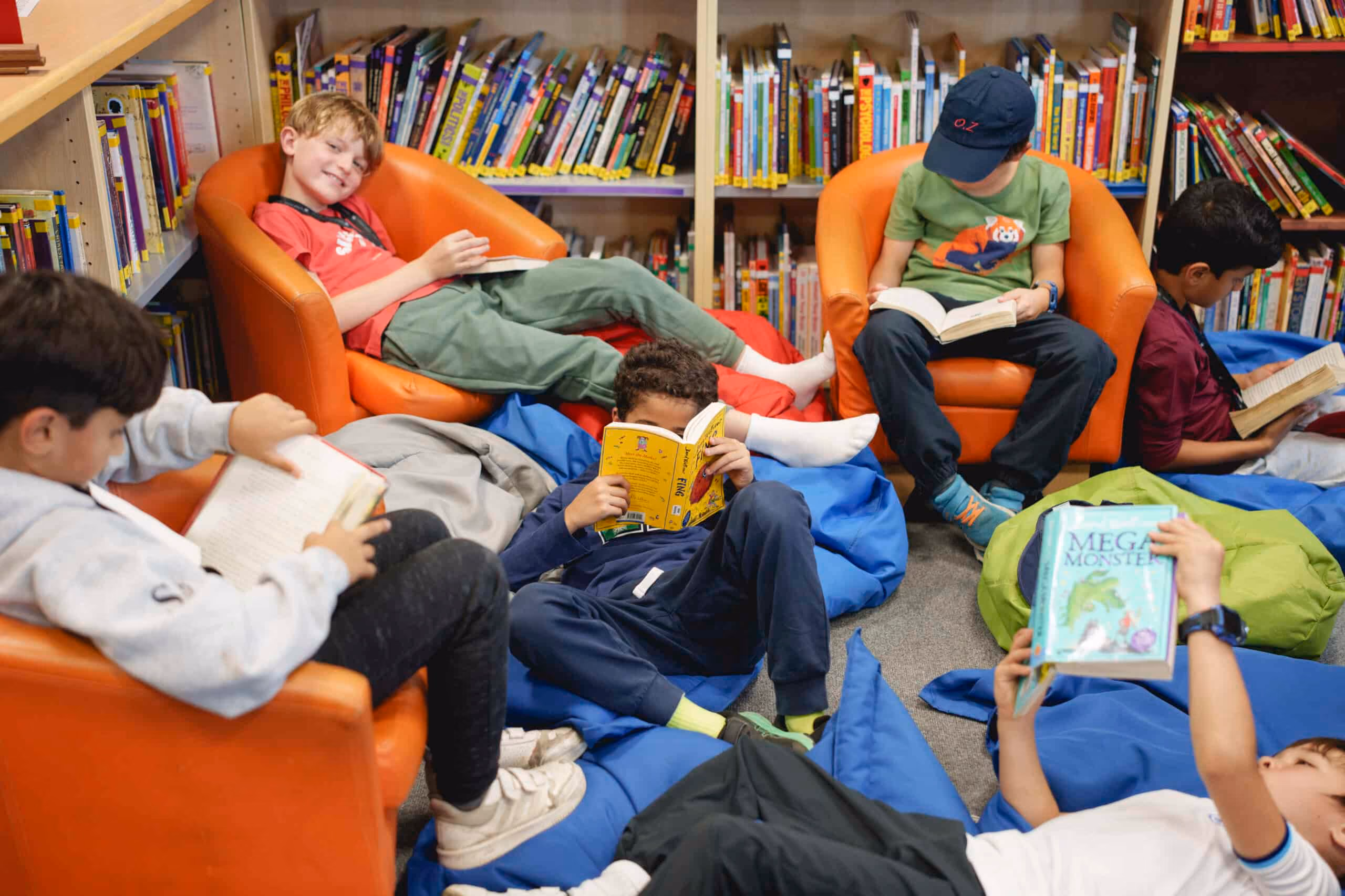 Children reading books in a library seated on orange chairs and colorful bean bags.