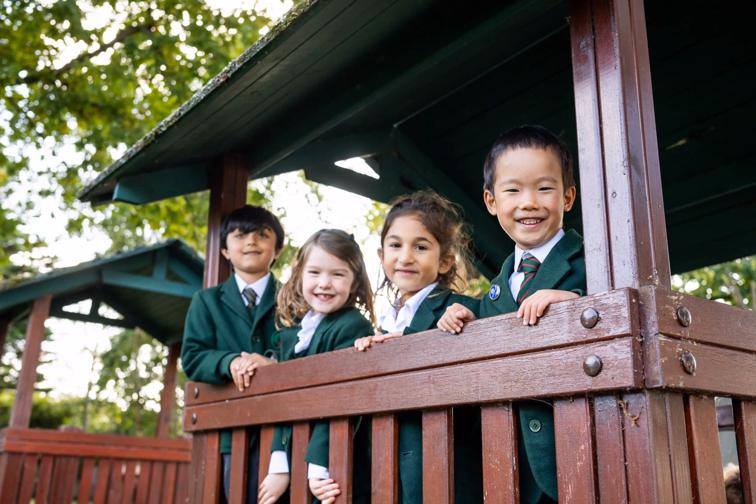 Four smiling children in green school uniforms standing on a wooden playhouse balcony outdoors.