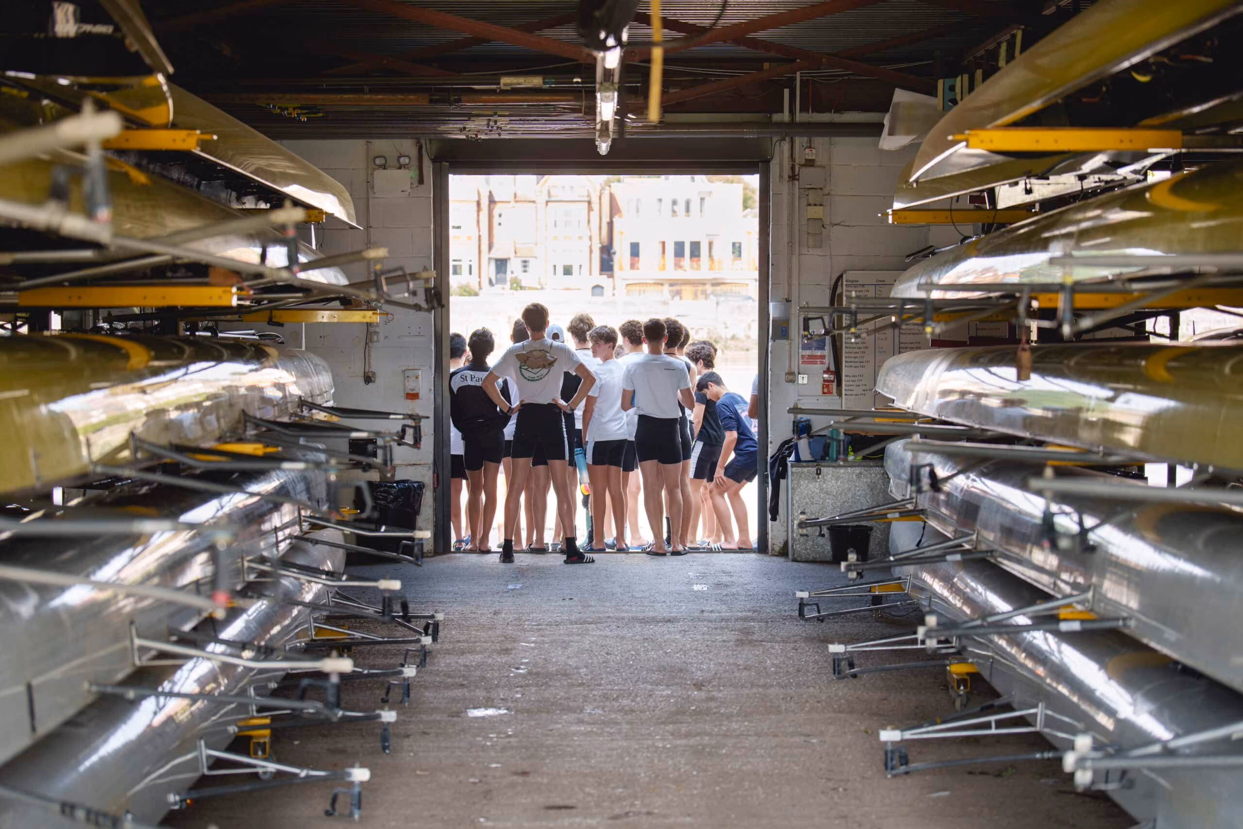 Group of rowers in shorts and t-shirts standing at a boathouse entrance with stacked rowing shells.