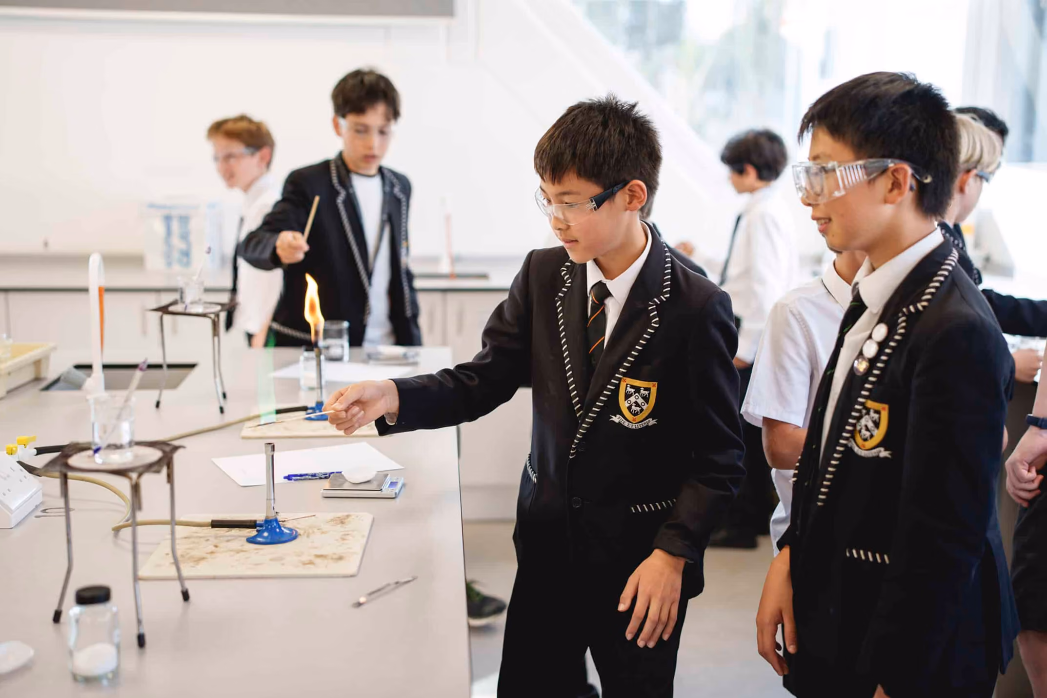 Middle school students in school uniforms and safety goggles conducting a chemistry experiment using Bunsen burners in a science lab.