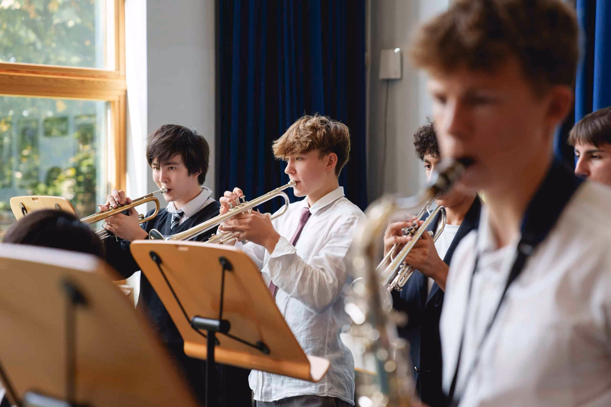 A group of teenage boys playing brass instruments, including trumpets and saxophone, in a music ensemble with sheet music stands in front of them.