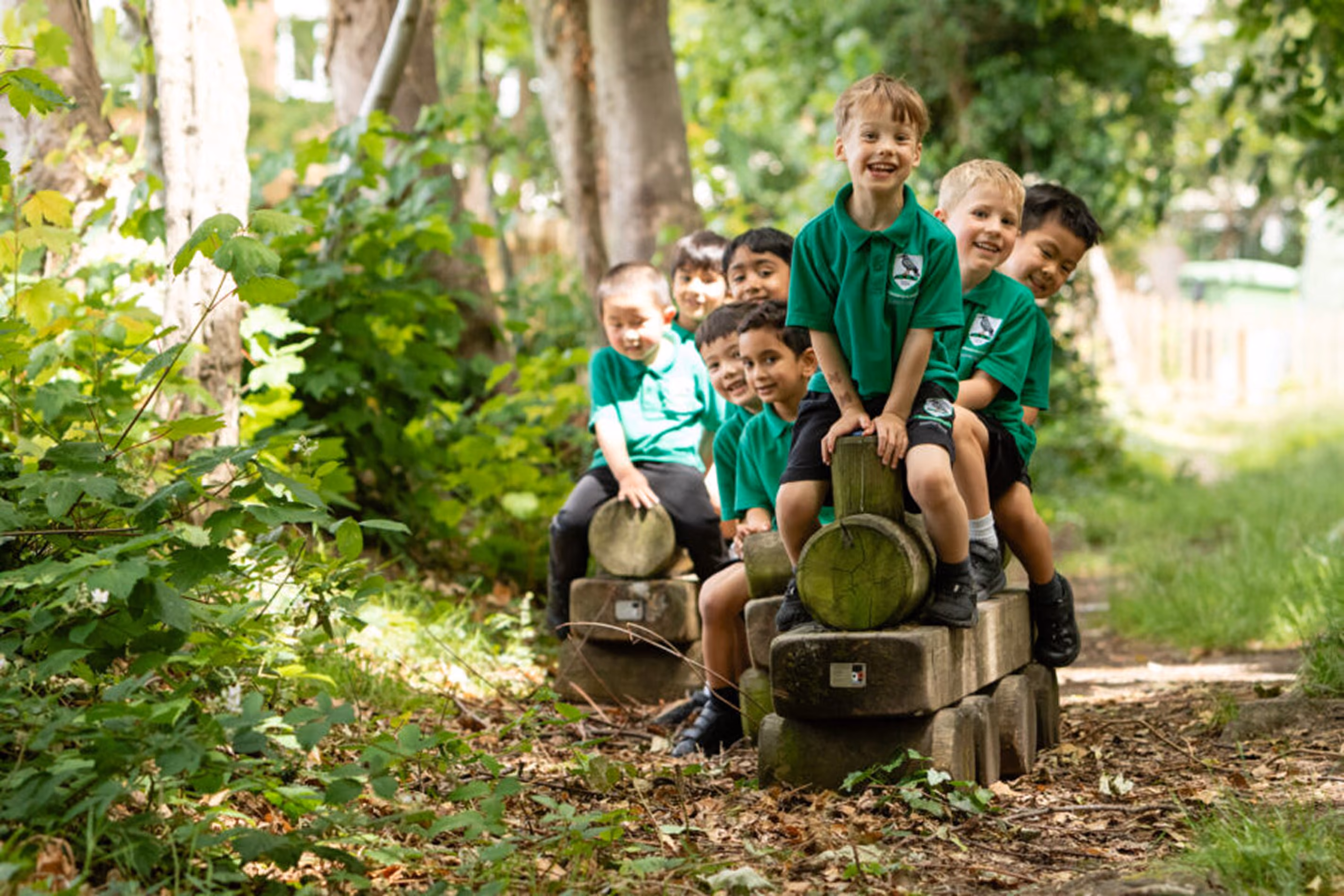 Group of young boys in green school uniforms sitting in a line on wooden play structures resembling a train outdoors surrounded by trees and greenery.