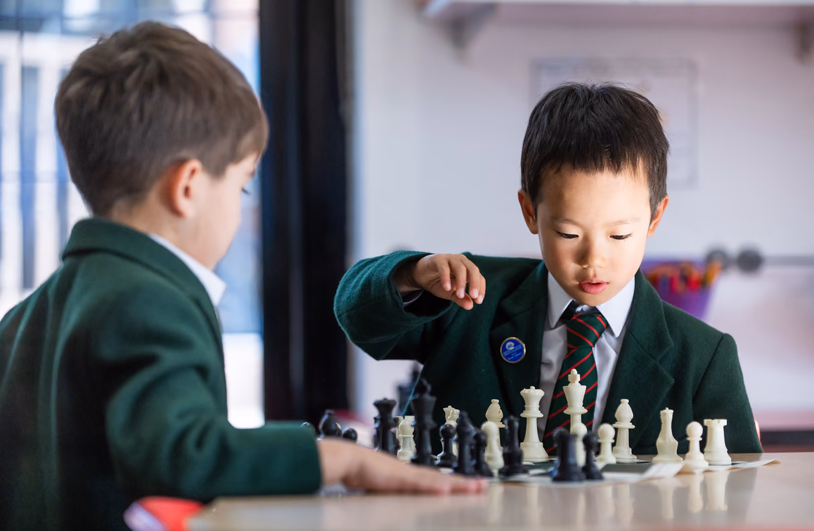 Two young boys in green school blazers play a game of chess at a table indoors, focused on the chessboard.