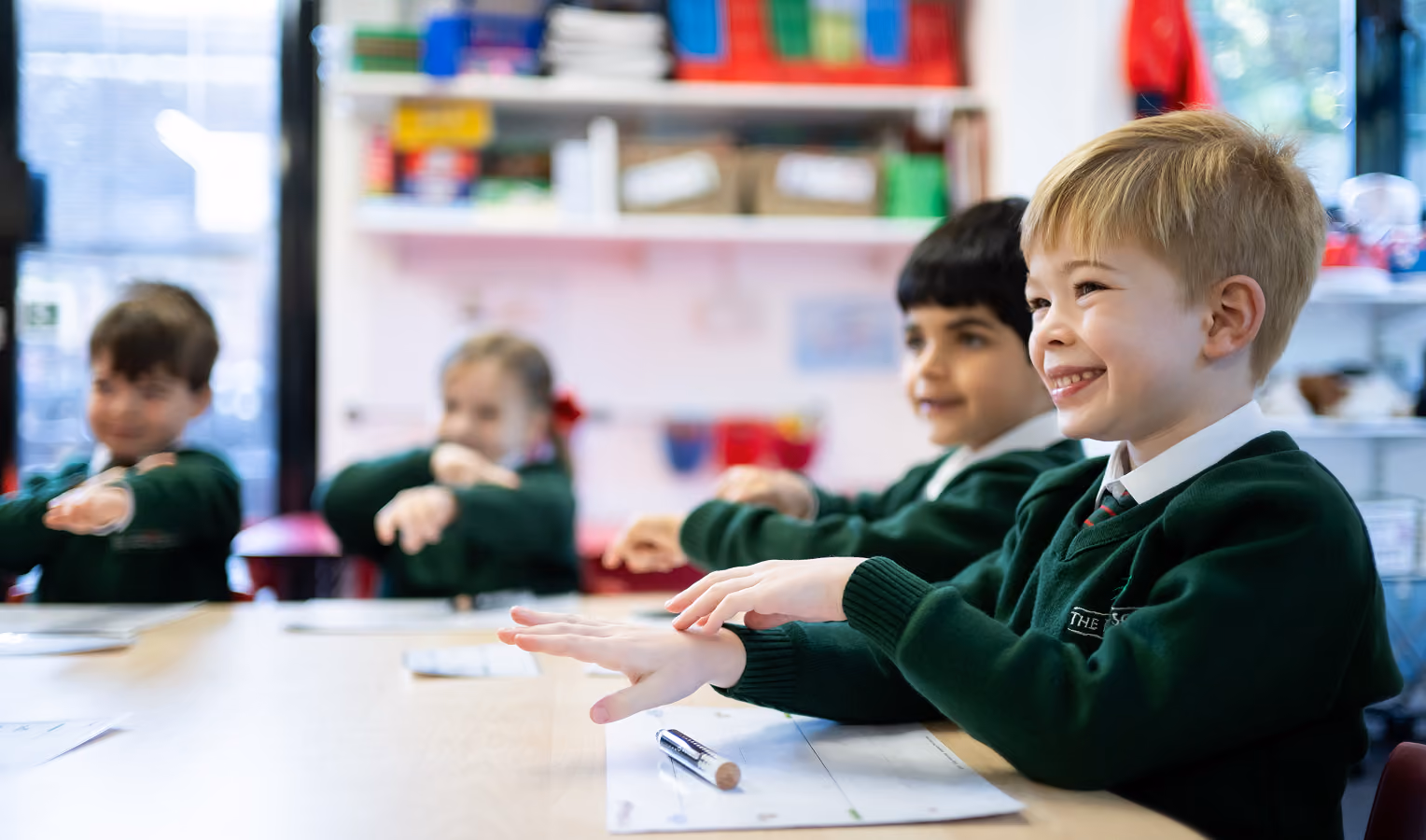 Four young children in green school uniforms sitting at a classroom table extending their arms forward, with one boy smiling brightly in the foreground.