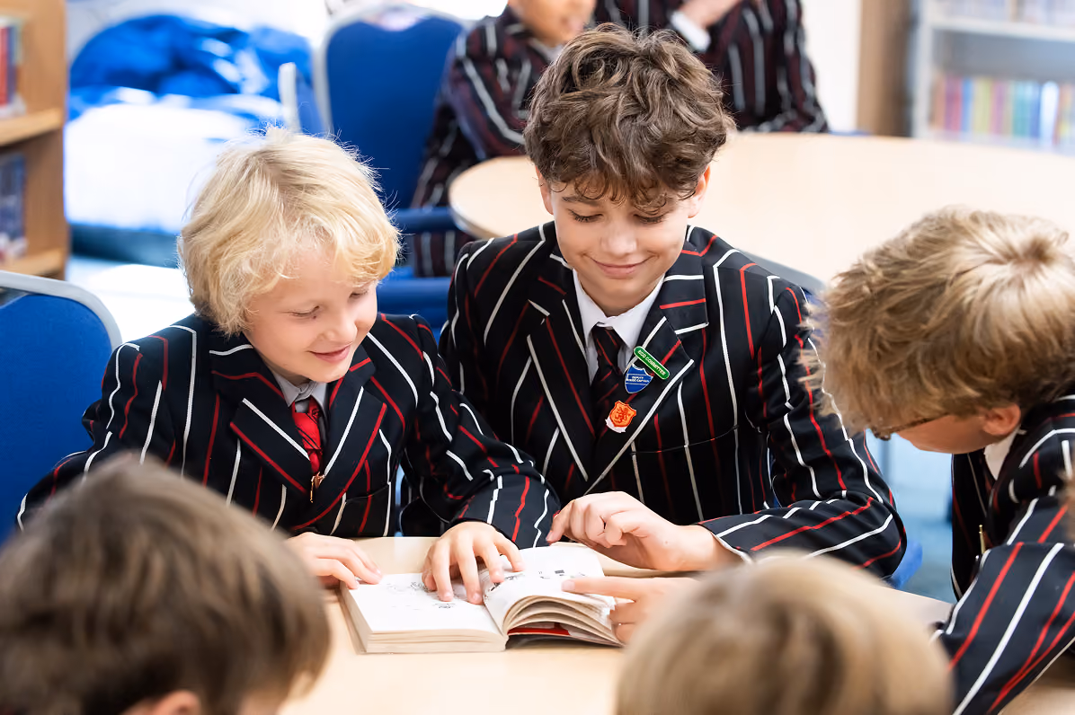 Four young boys in black blazers with red and white stripes and red ties sit around a table in a classroom, engaged with an open book that one boy is pointing at while others look on with interest.