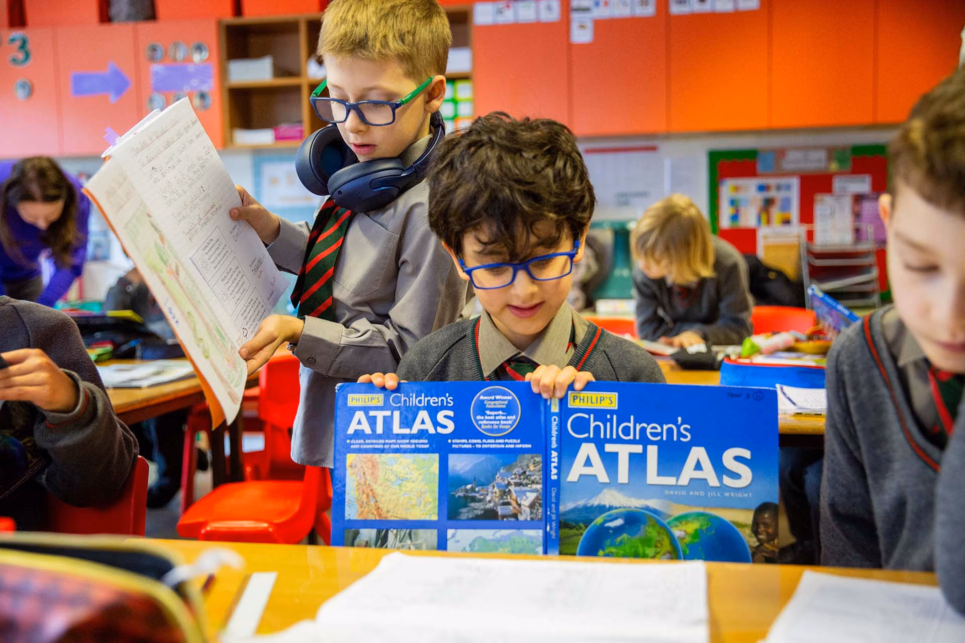 School children in uniform reading Philip's Children's Atlas books at a classroom table with colorful shelves and educational materials in the background.