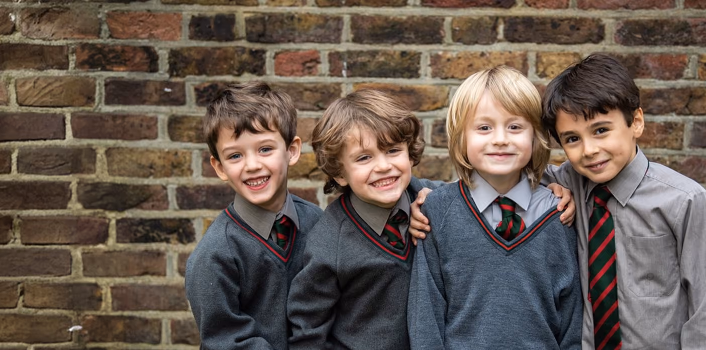 Four young boys standing close together in front of a brick wall, smiling and wearing school uniforms with grey sweaters and striped ties.