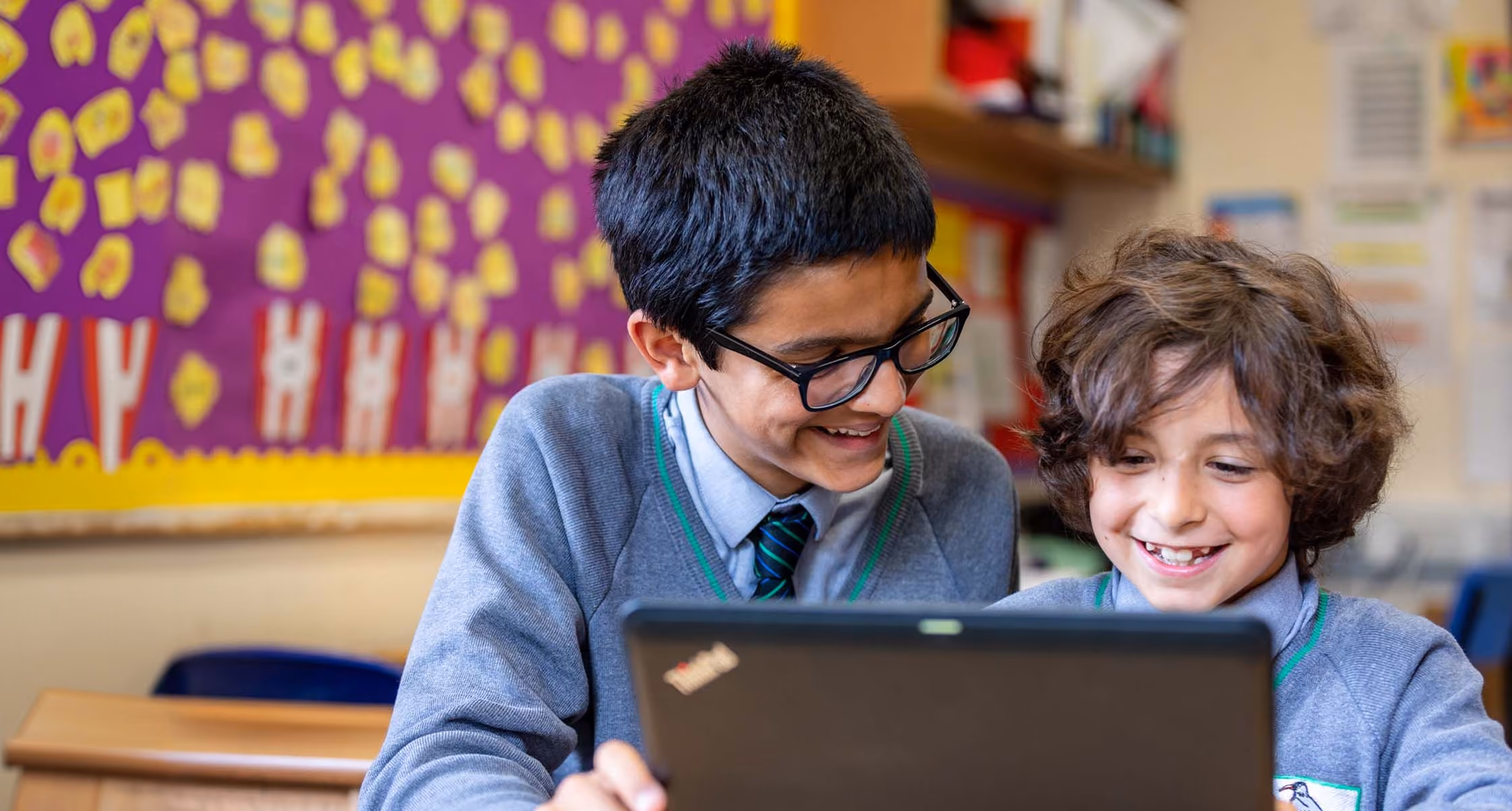 Two schoolboys in grey uniforms sitting at a desk and looking at a Lenovo ThinkPad laptop, smiling in a classroom with colorful bulletin board in the background.