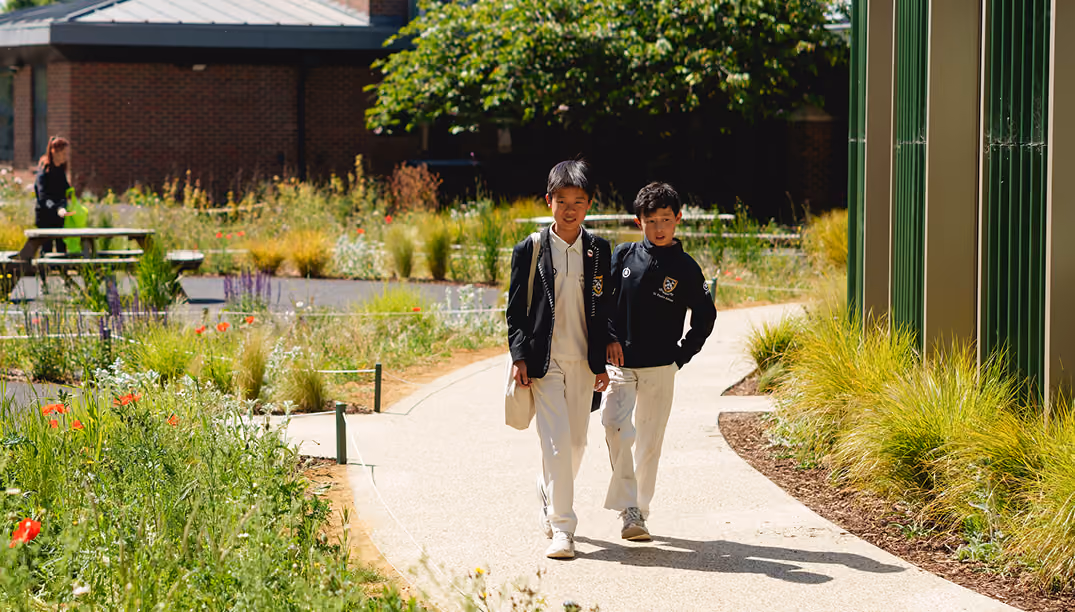 Two schoolboys in uniforms walking on a garden path surrounded by greenery and flowers, with a picnic table and a brick building in the background.