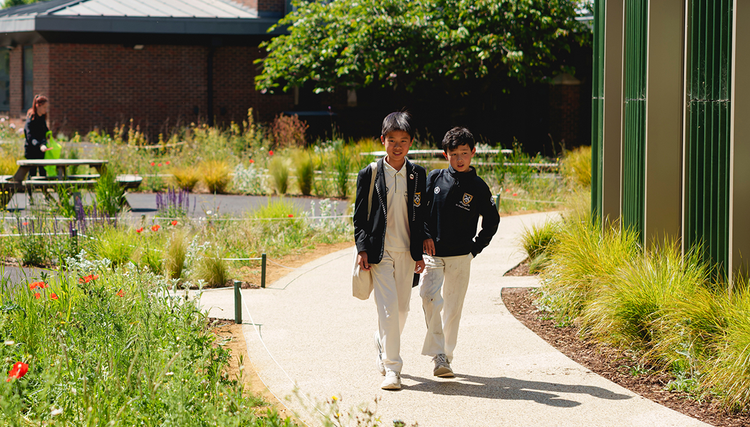 Two schoolboys in uniforms walking on a garden path surrounded by greenery and flowers, with a picnic table and a brick building in the background.