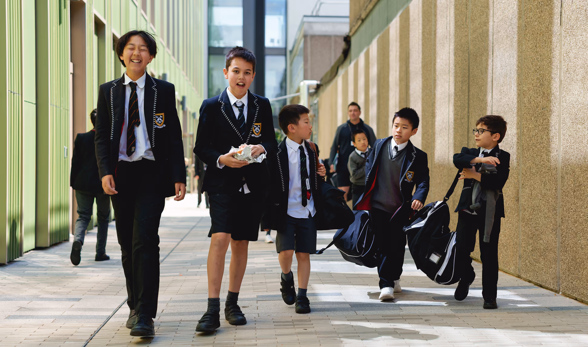 A group of five boys in school uniforms walking outside on a paved path between buildings, carrying bags and smiling.