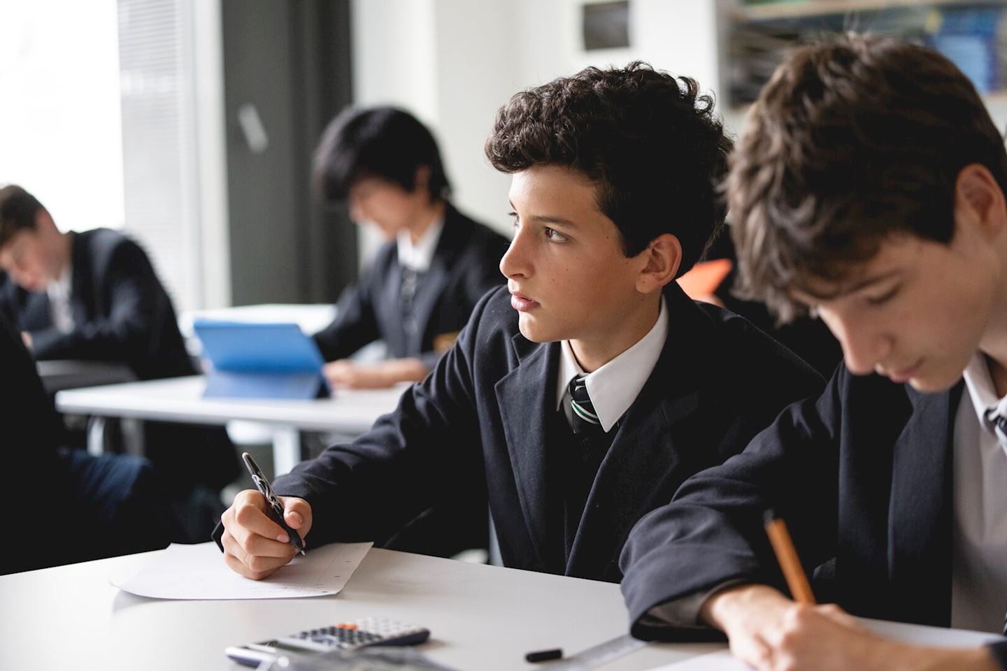 Teenage boys in school uniforms sitting at desks in a classroom, writing on papers, with a calculator and pencil case on the table, one boy looking away thoughtfully.