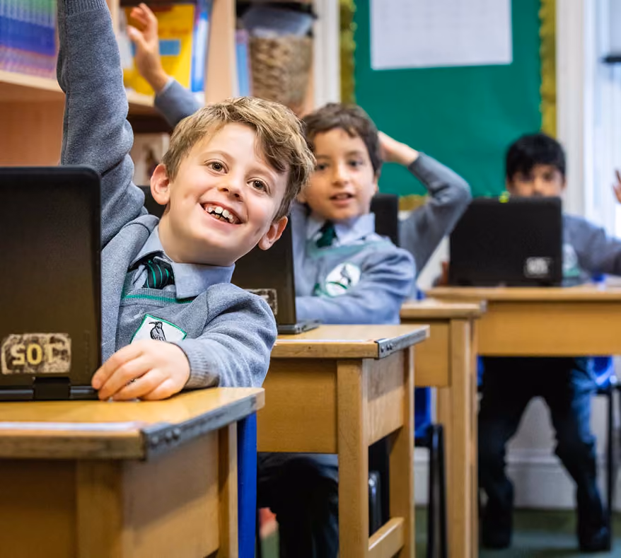 Three schoolboys in grey uniforms sitting at wooden desks, each with a laptop; the boy closest to the camera is smiling and raising his hand.
