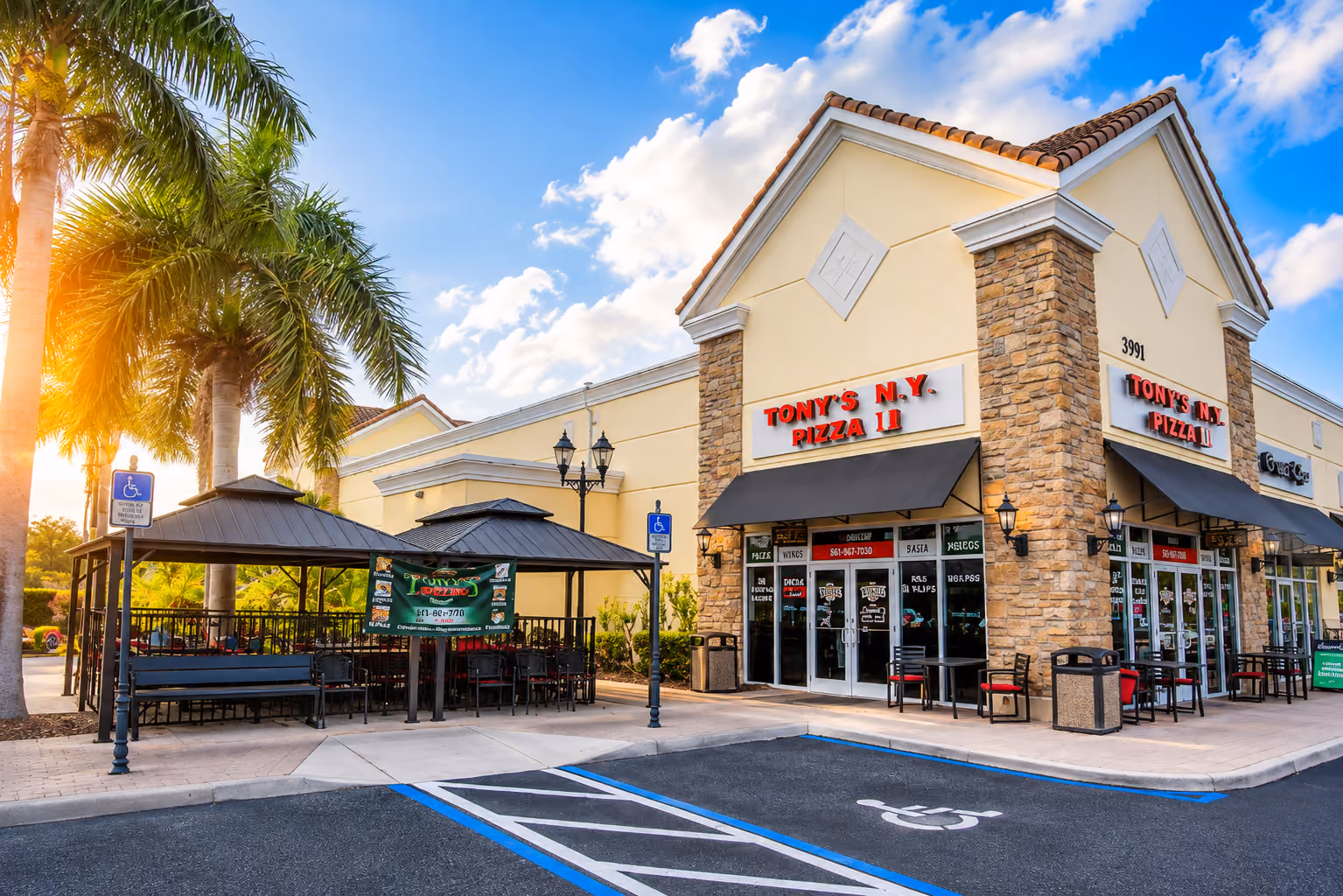 Outdoor seating area and entrance of Tony's N.Y. Pizza II under blue sky with palm trees.