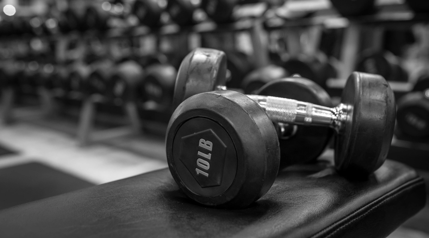 A close-up black-and-white photograph of a 10 LB dumbbell lying on a dark padded gym bench with rows of weight racks blurred in the background.