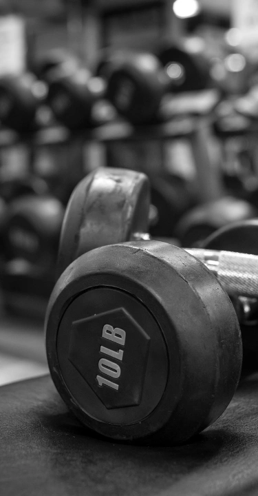 A close-up black-and-white photograph of a 10 LB dumbbell lying on a dark padded gym bench with rows of weight racks blurred in the background.
