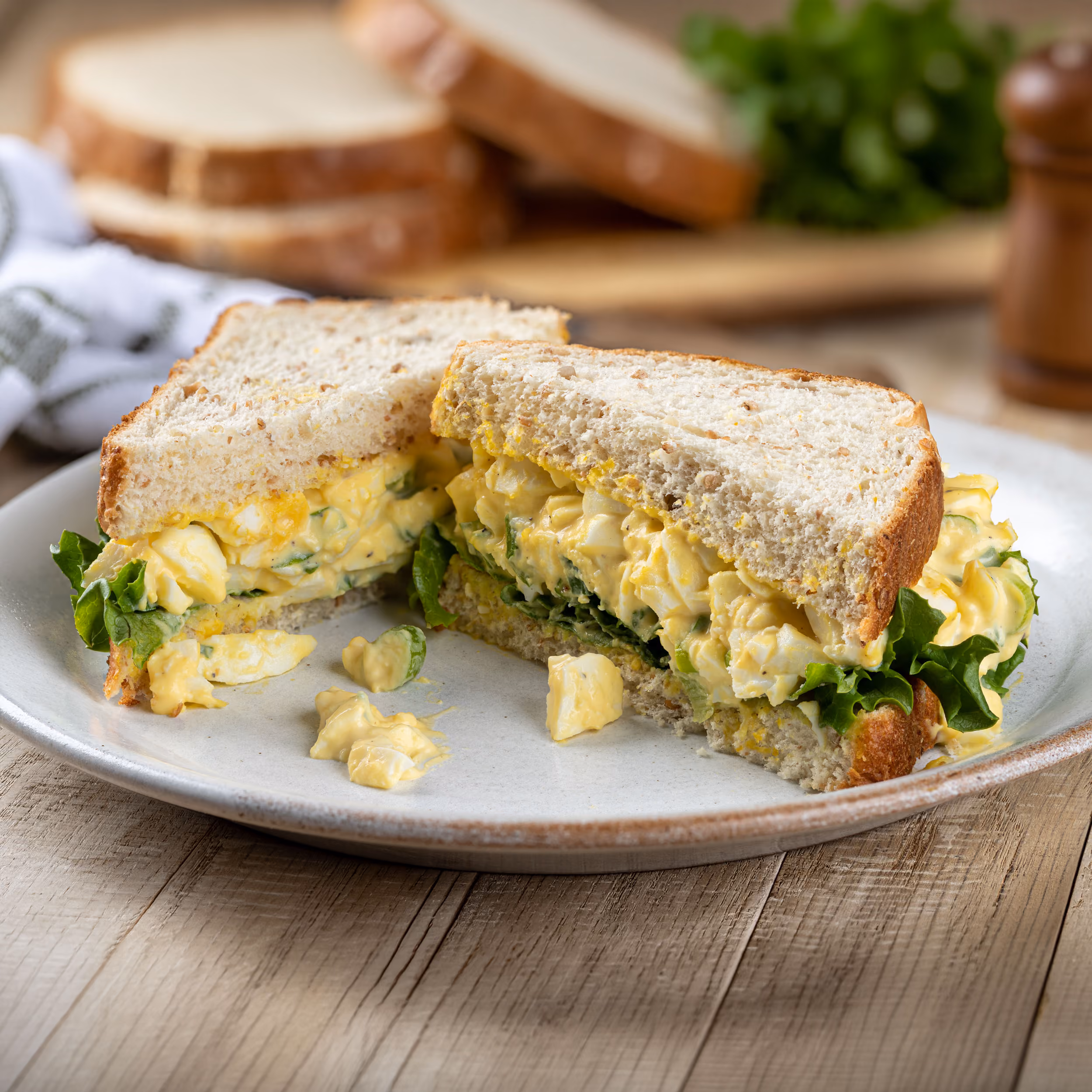 Egg sandwich halves with lettuce on whole wheat bread on a white plate on a wooden table.