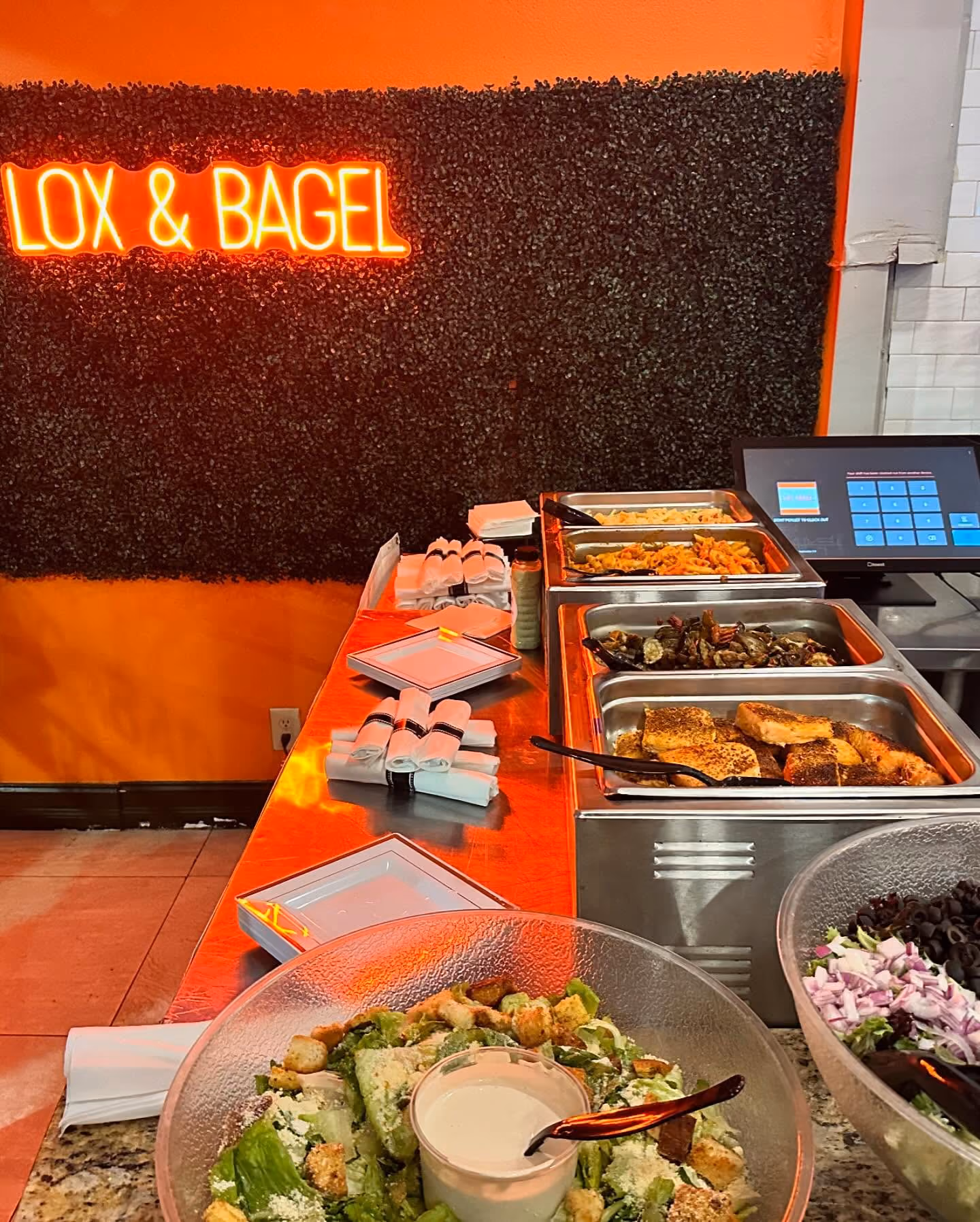 Buffet table with trays of food, salads, napkins, and a neon sign reading LOX & BAGEL.