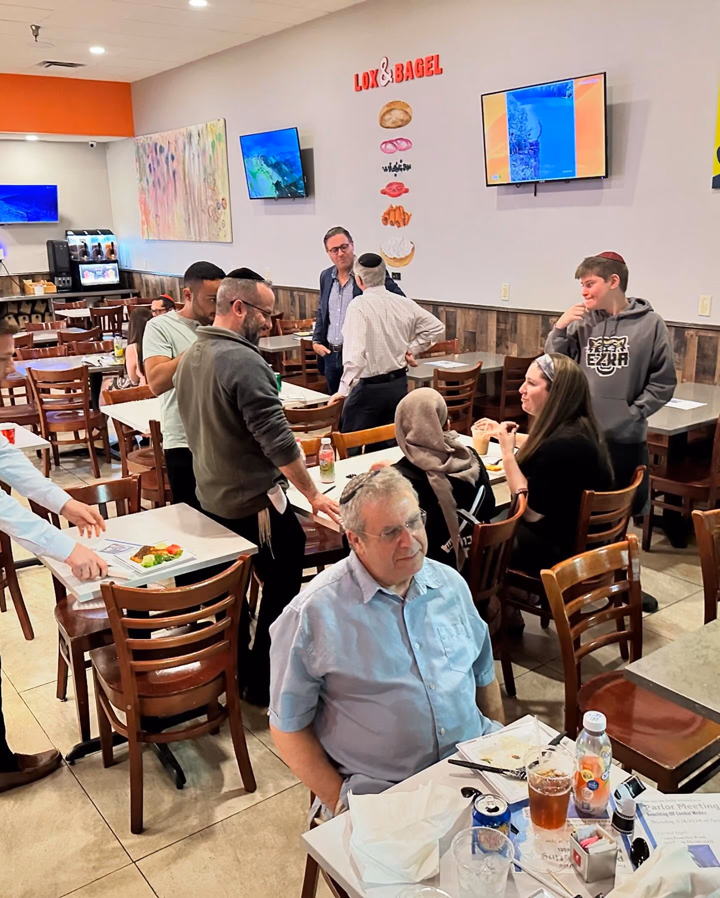 People dining and conversing inside Lox & Bagel restaurant with wall art and TVs.