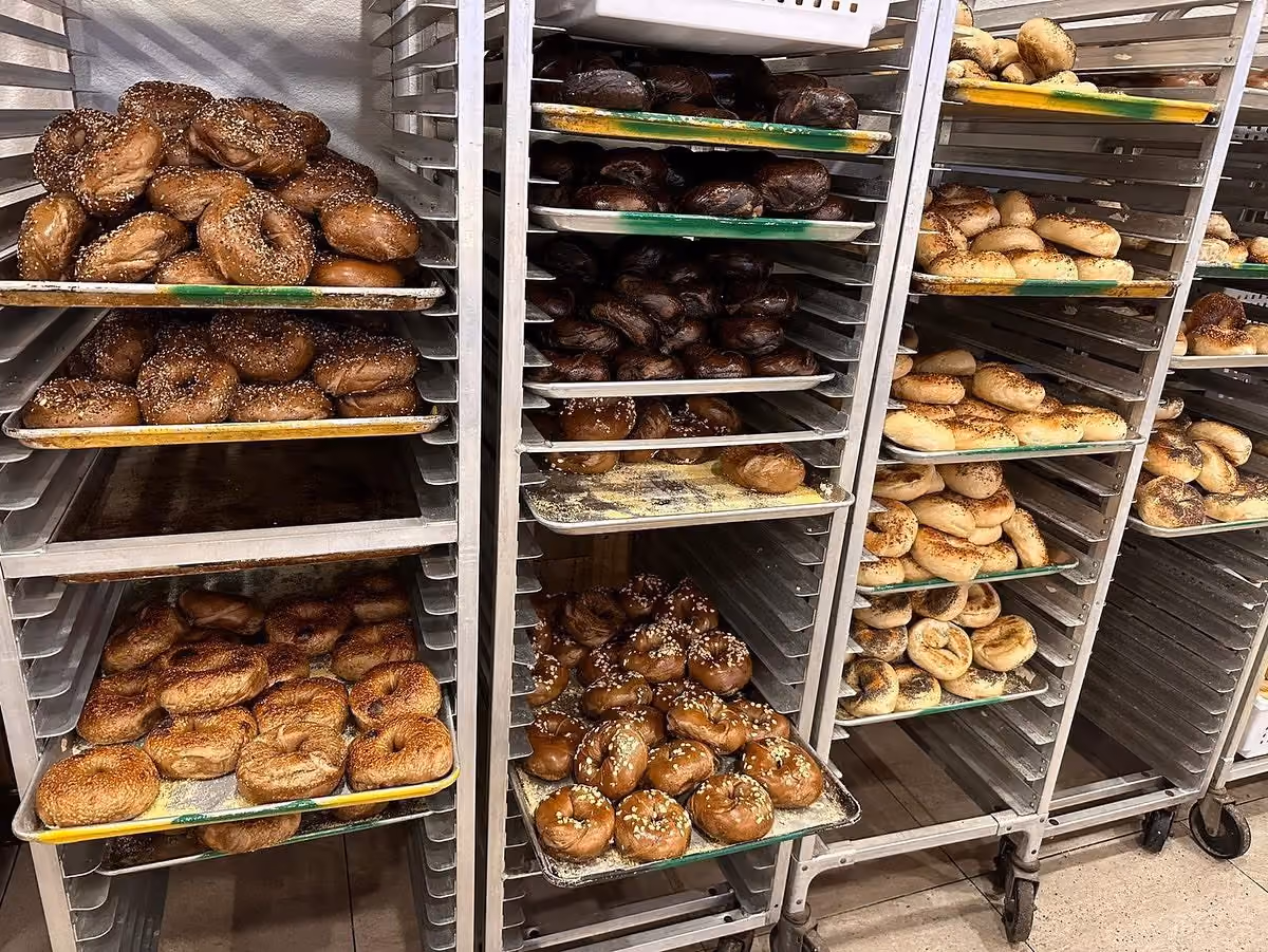 Metal racks filled with trays of various bagels including plain, seeded, and dark bagels.