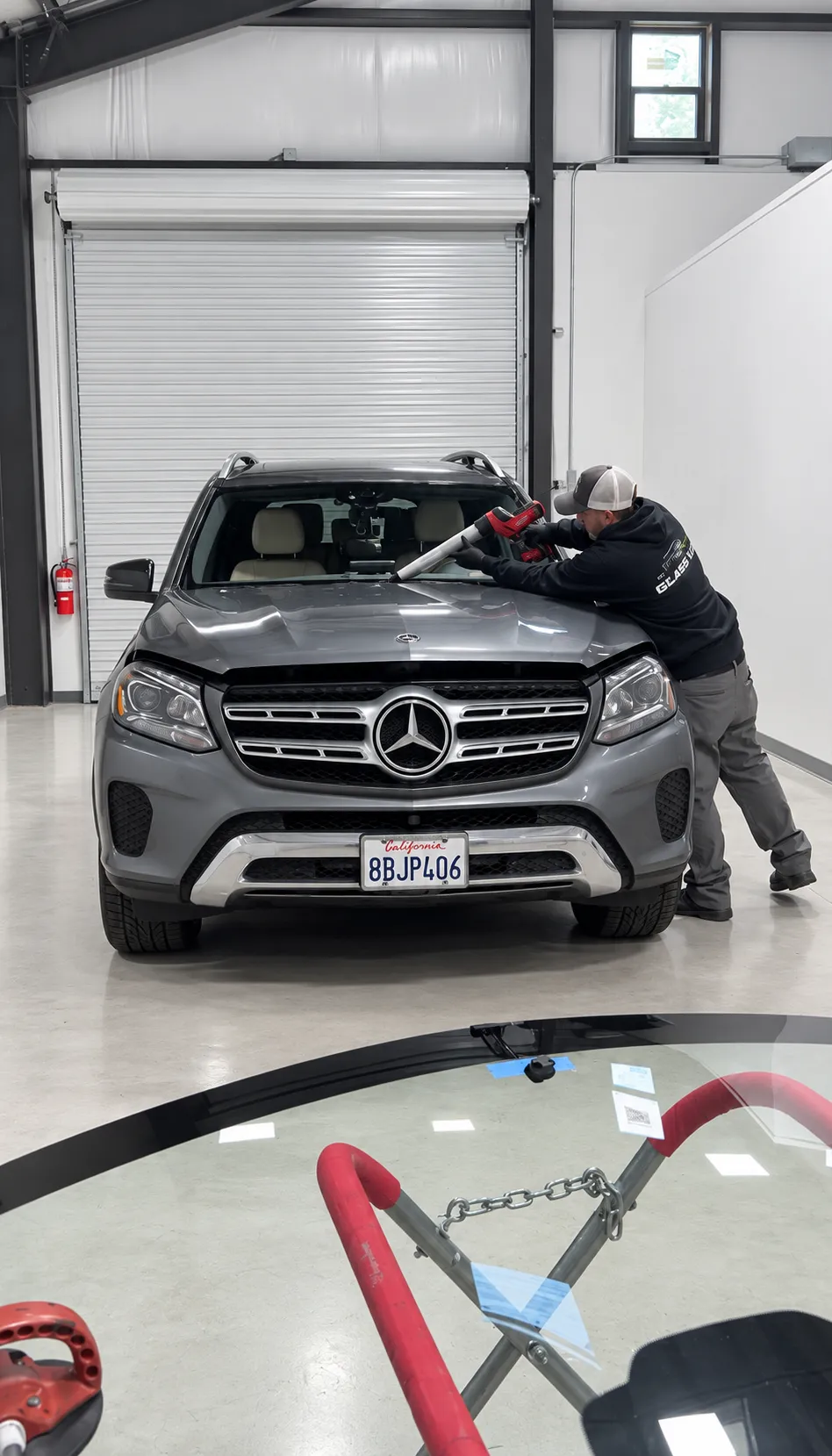 Technician working on a gray Mercedes-Benz SUV windshield, with glass on a stand in the foreground.