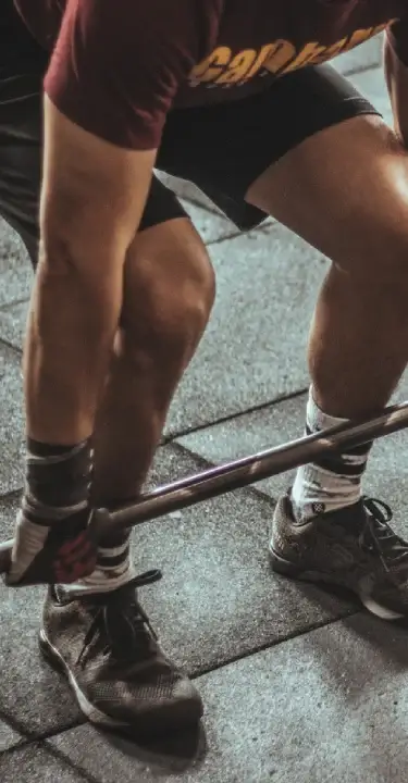 Close-up of an athlete bending down to lift a barbell in a gym, wearing shorts, training shoes, and wrist wraps on a rubber floor.