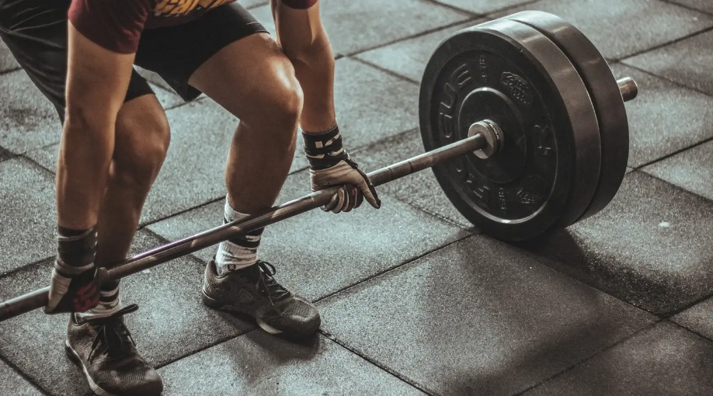 Close-up of an athlete bending down to lift a barbell in a gym, wearing shorts, training shoes, and wrist wraps on a rubber floor.