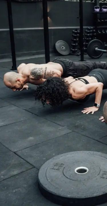 Three athletes performing push-ups together on a gym floor, surrounded by barbells, dumbbells, weight plates, and other strength-training equipment.