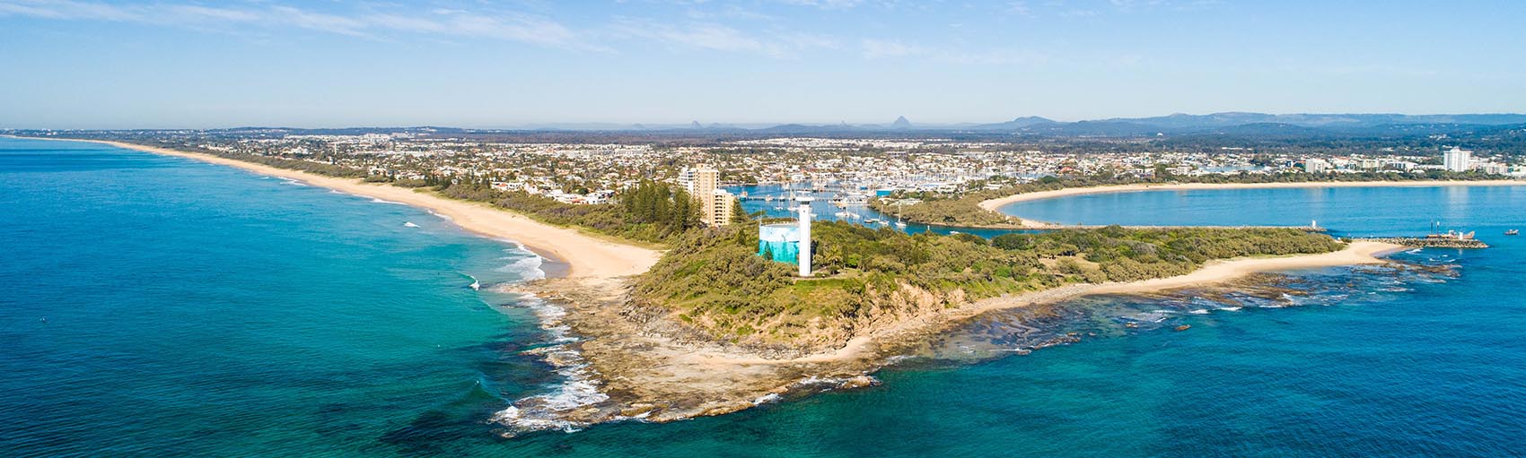 Aerial view of a coastal peninsula with a white lighthouse surrounded by greenery, sandy beaches stretching on both sides, blue ocean waters, and a town in the background under a partly cloudy sky.