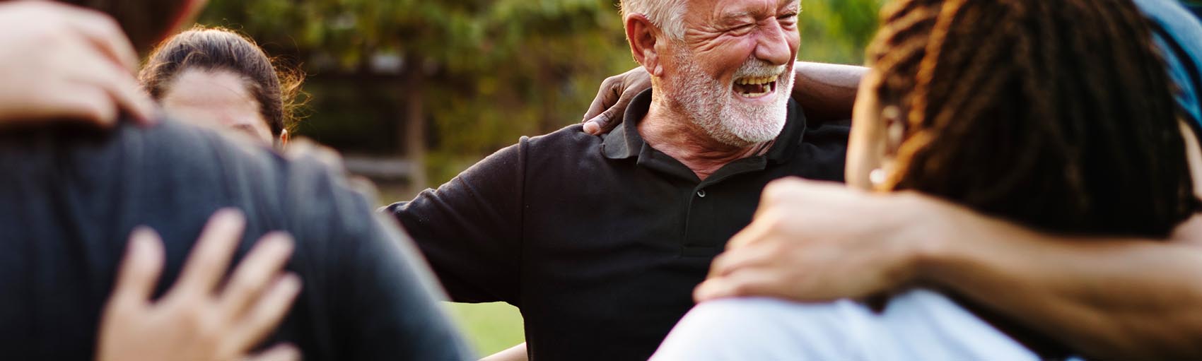 Older man with white hair and beard smiling and embracing a diverse group of people in an outdoor setting.