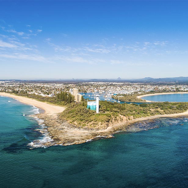 Aerial view of a coastal town featuring a small peninsula with a lighthouse, surrounded by clear blue ocean, sandy beaches, green vegetation, and a harbor with boats docked.