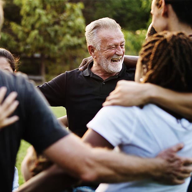 Smiling older man with gray hair and beard wearing a black shirt embraces a group of people outdoors in a park setting.