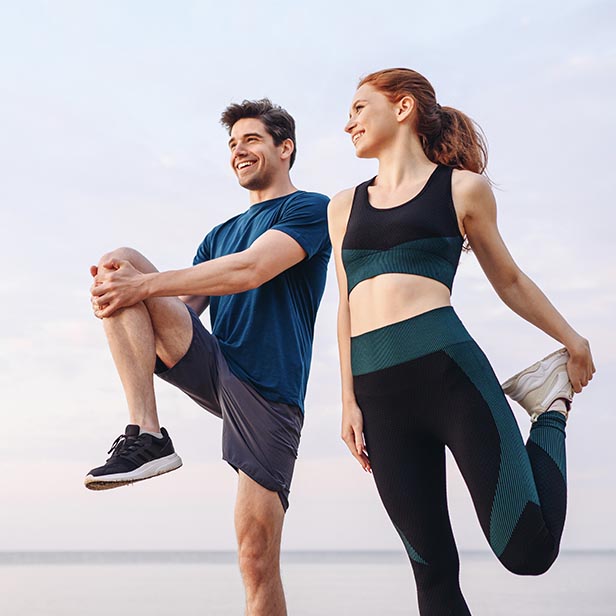 A smiling man and woman in athletic wear are stretching outdoors against a calm sky, preparing for exercise or running.