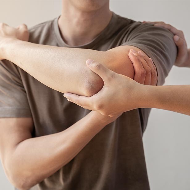 A person in a brown shirt is holding their bent elbow while another person supports the arm near the shoulder and forearm, suggesting elbow massage therapy or joint support.