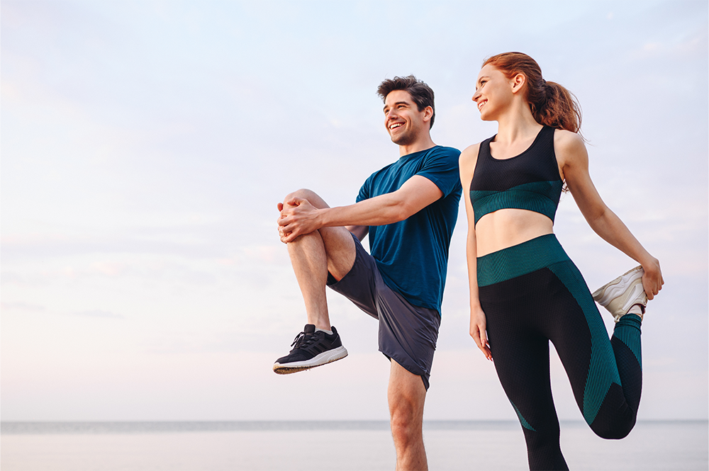 A man and woman outdoors near water performing leg stretches, smiling and wearing athletic clothing, under a partly cloudy sky.