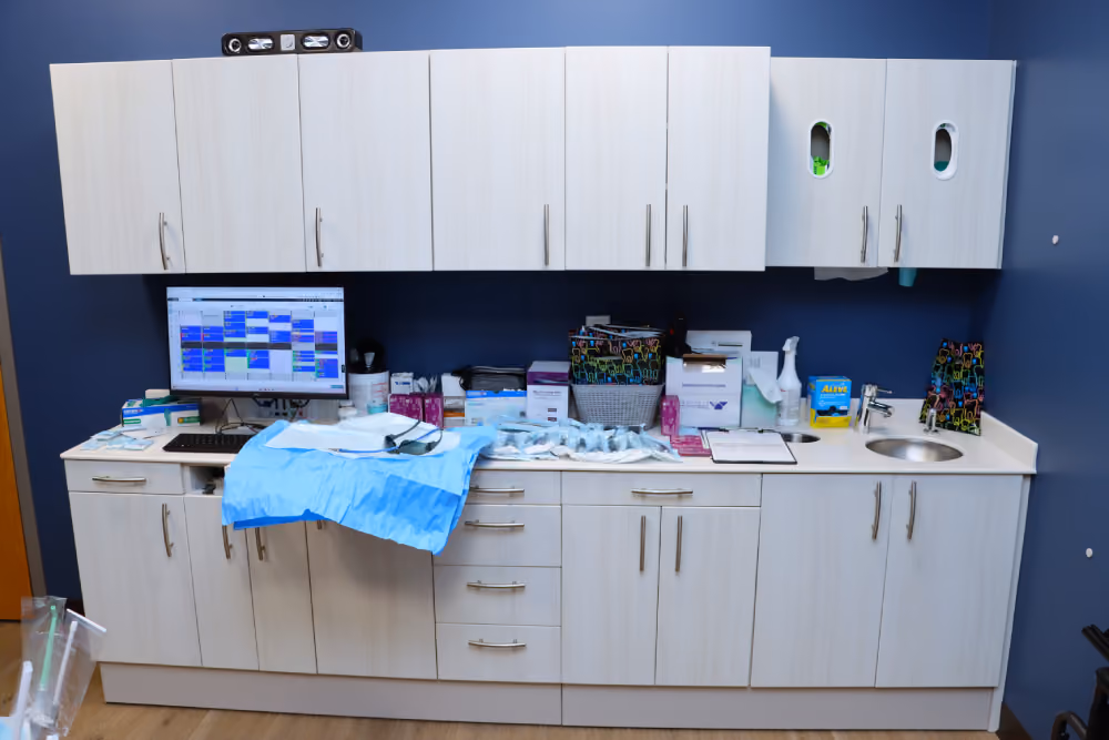Clean and organized dental or medical workspace with white cabinets and a countertop holding supplies including gloves, dental bibs, paperwork, and a computer screen displaying a schedule against a blue wall.
