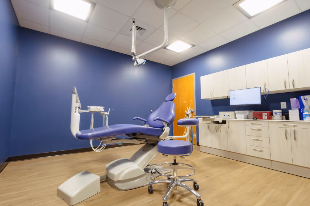 Modern dental examination room with a purple dental chair and matching stool in the center, blue walls, wood flooring, overhead lighting, and light-colored cabinets with a monitor and dental equipment.