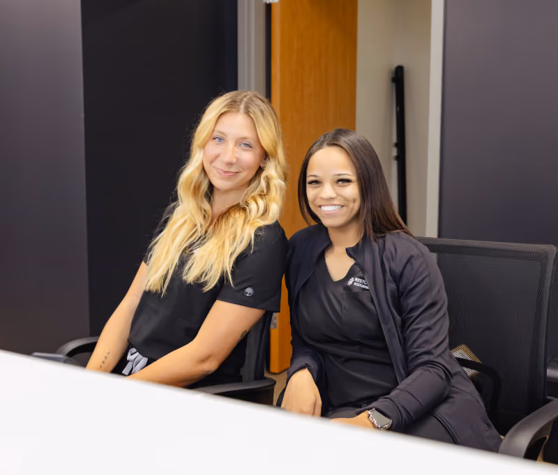 Two women sitting side by side on black office chairs behind a white table, smiling and looking at the camera, with a dark background and a partially open wooden door behind them.