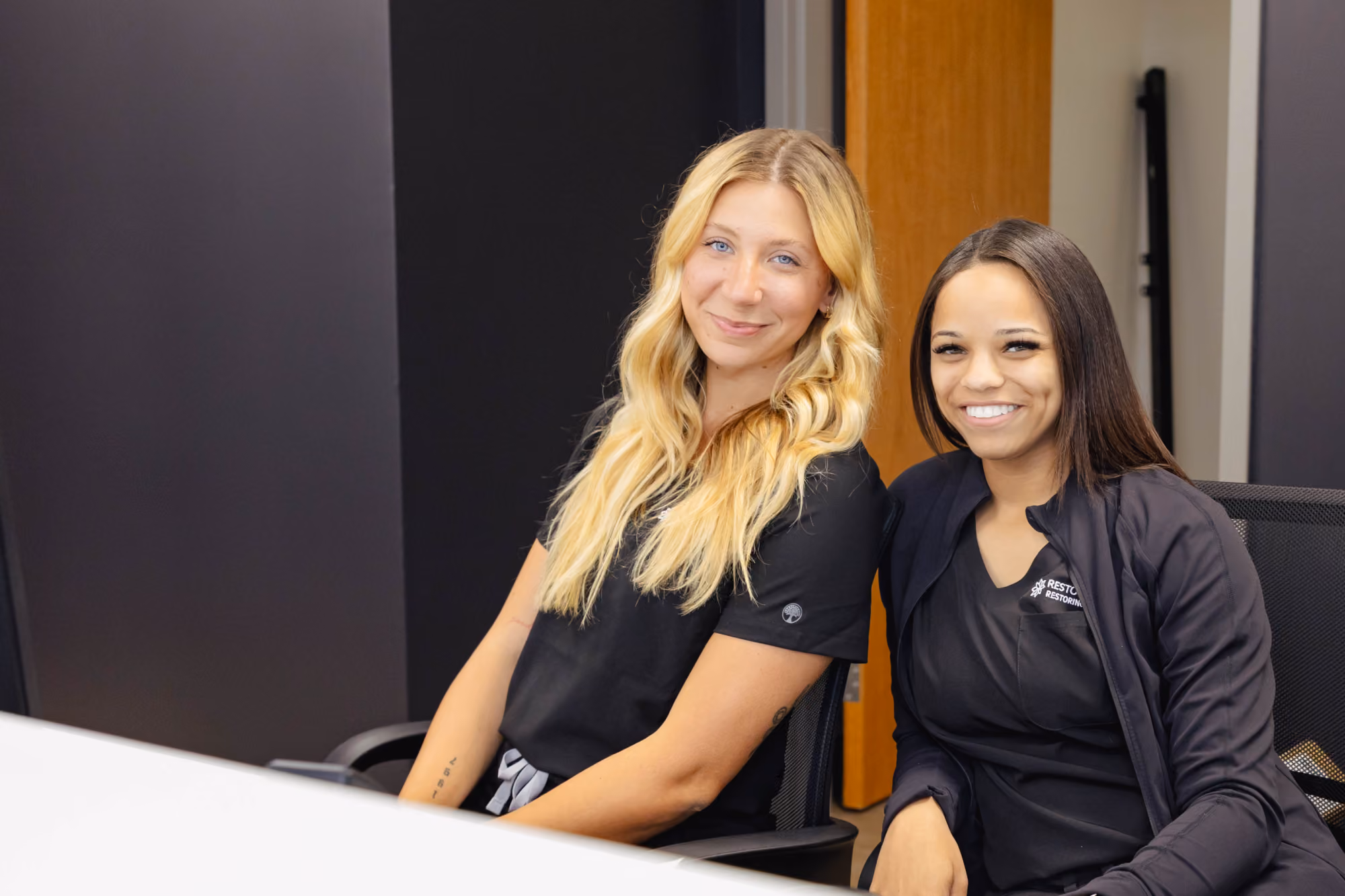 Two women sitting side by side in office chairs, smiling at the camera; the woman on the left has long blonde hair and wears a black shirt, the woman on the right has straight dark hair and wears a black jacket over a black top.