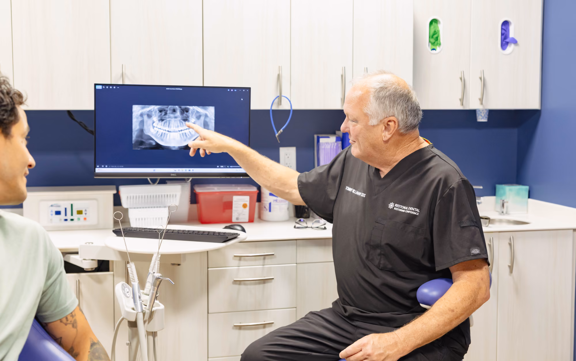 Dentist pointing at a dental X-ray on a computer screen while talking to a seated patient in a dental office.