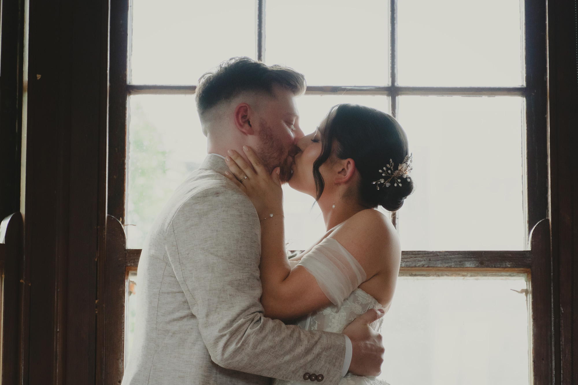 Bride and groom sharing a kiss in front of a large window on their wedding day.